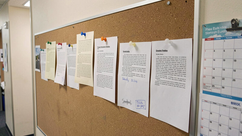 A row of dated printed notices pinned to a corkboard in an office corridor, with a wall calendar beside them.