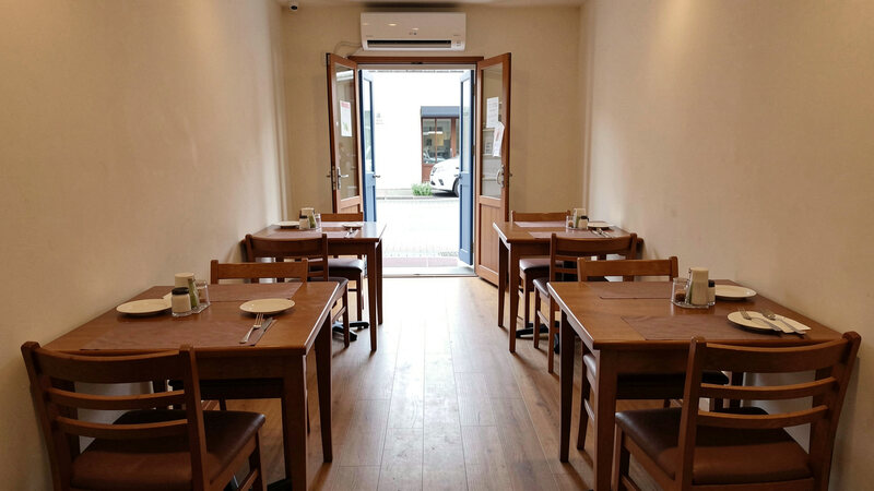 A small restaurant dining room with tables spaced apart, chairs tucked in and a window open at the far end.