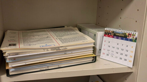 A stack of dated printed notices and a closed ring binder on a shelf, with a wall calendar open beside them.