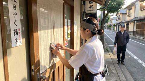 A small Japanese shop entrance in the morning, a staff member wiping the door handle with a cloth while a customer waits on the pavement outside.