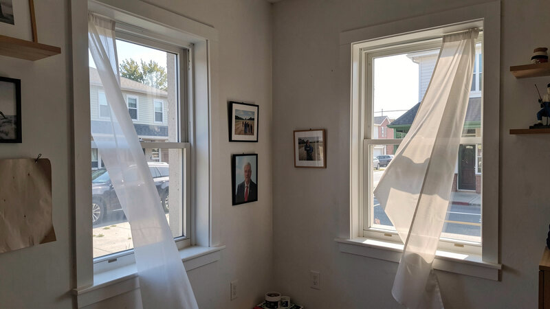 Two windows open on opposite walls of a small shop, with a light curtain moving in the draught and daylight entering.